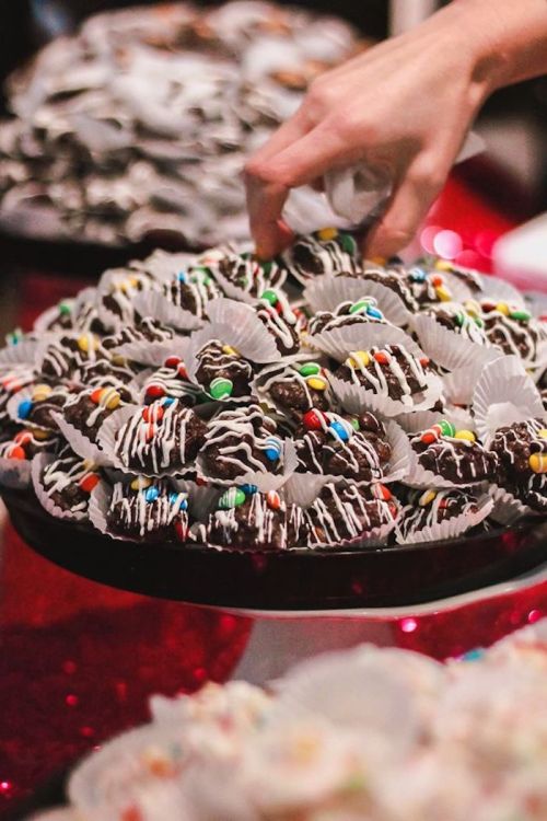 A hand reaches for a plate of chocolate-covered treats decorated with colorful sprinkles and drizzled with white icing.