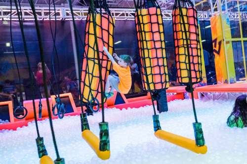A child climbs on colorful hanging obstacles above a ball pit in a vibrant indoor play area.