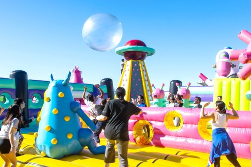 A vibrant inflatable playground with people playing and a large beach ball in the air under a clear blue sky.