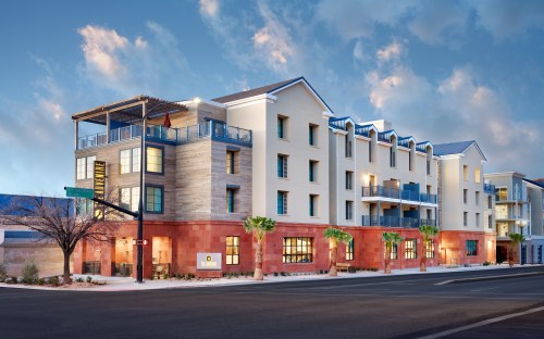 Modern hotel building with a mix of stone and wood exterior, featuring balconies and palm trees, under a blue sky.