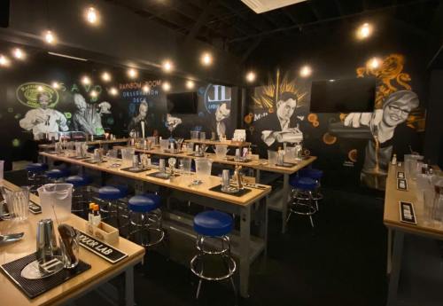 A vibrant bar setting with tables, stools, and colorful murals of bartenders on the walls, illuminated by hanging lights.