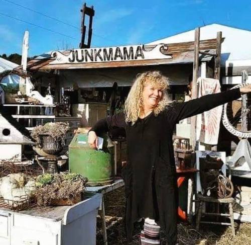 A smiling woman with curly hair stands in front of a vintage market stall labeled "JunkMama," surrounded by eclectic items.
