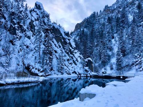 Snow-covered mountains and trees reflect in a calm, icy blue lake under a cloudy sky.