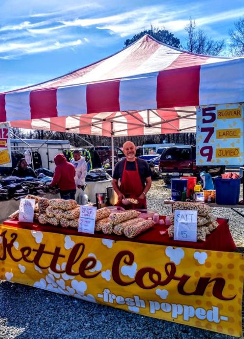 A vendor stands behind a kettle corn stall with bags of popcorn under a red and white striped tent at a market.