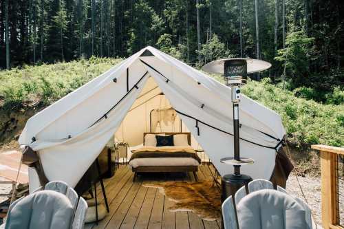 A cozy glamping tent with a bed, wooden deck, and outdoor heater, surrounded by lush greenery and tall trees.