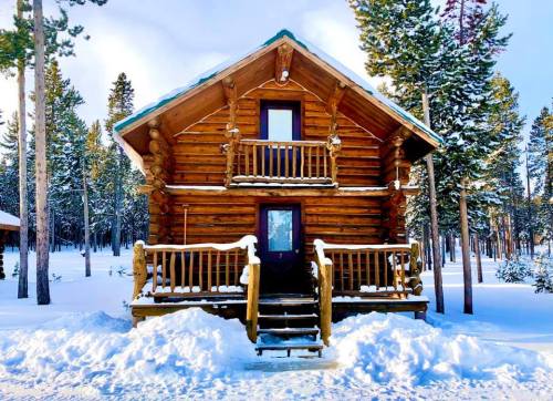 A cozy log cabin surrounded by snow-covered trees, featuring a front porch and two balconies.
