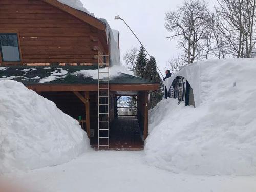 A person uses a long pole to remove snow from a roof, surrounded by large snowdrifts near a wooden house.