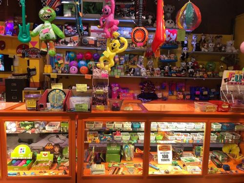 Colorful arcade prize counter filled with toys, games, and collectibles, showcasing various items behind glass displays.