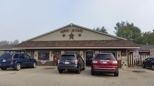 A cozy café with a sign reading "Curve-It Café," featuring parked cars and patriotic decorations outside.