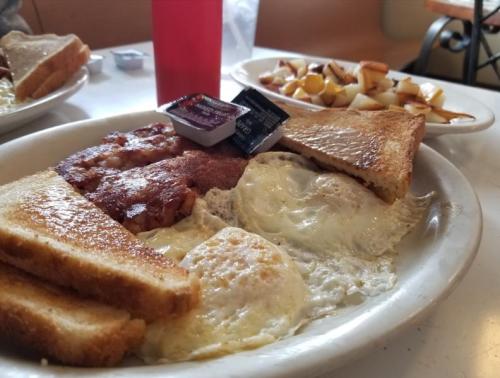 A plate of breakfast featuring eggs, toast, bacon, and a side of diced potatoes with condiments.