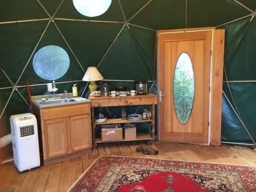 Interior of a geodesic dome with a wooden door, kitchenette, and a red rug on the wooden floor.