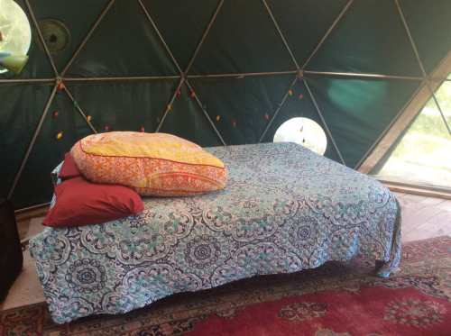 Cozy interior of a geodesic dome with a bed covered in a colorful blanket and decorative pillows. Natural light filters in.