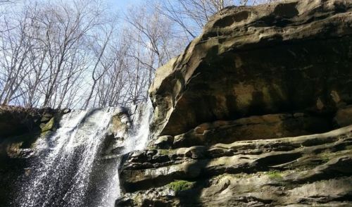 A waterfall cascading over rocky cliffs, surrounded by bare trees under a clear blue sky.