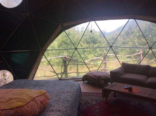 Interior of a geodesic dome with a bed and couch, featuring large windows overlooking a green landscape.