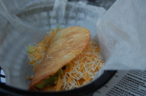 A crispy taco shell filled with shredded cheese and lettuce, placed in a basket lined with paper.