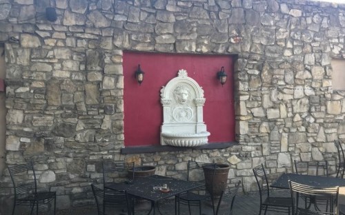 A stone wall with a red section featuring a decorative white fountain and outdoor tables and chairs in front.