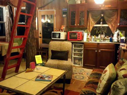 Cozy cabin interior with a red ladder, kitchen area, and eclectic decor, featuring a leopard-print chair and colorful rugs.