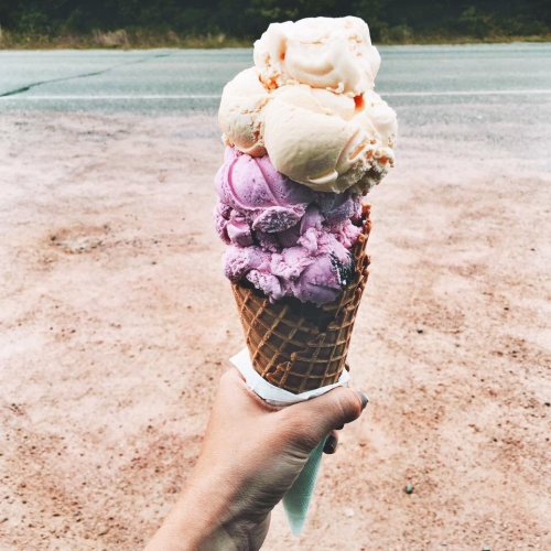 A hand holding a tall ice cream cone with three scoops: purple, orange, and white, against a gravel background.