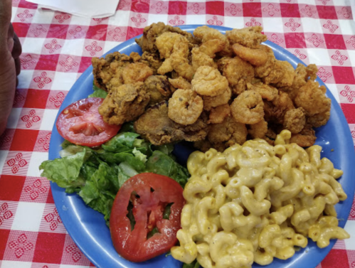 A blue plate with fried shrimp, lettuce, tomato slices, and macaroni and cheese on a red checkered tablecloth.