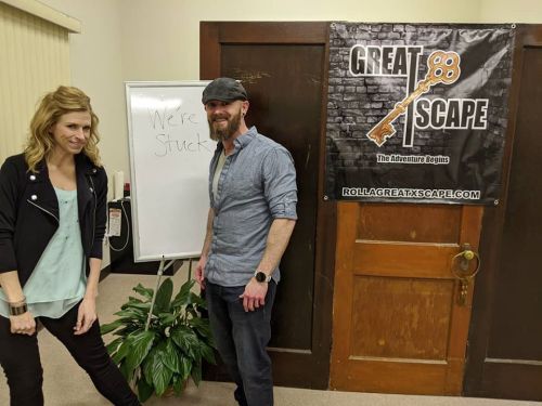 A man and woman stand next to a whiteboard that says "We're Stuck" in front of a Great Escape banner.