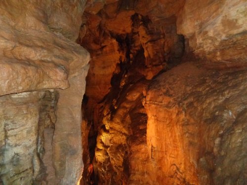 A narrow cave passage with textured rock walls illuminated by warm lighting, revealing stalactites and stalagmites.