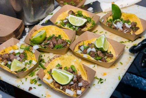 Six tacos filled with beef, onions, and cilantro, garnished with lime wedges, served in brown paper trays.