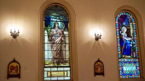 Two stained glass windows in a church, featuring a figure in a robe and another woman holding flowers, illuminated by wall sconces.