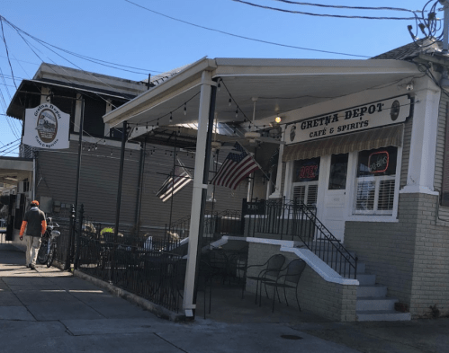 A café with a covered porch, flags displayed, and a sign that says "Open," set in a sunny street scene.