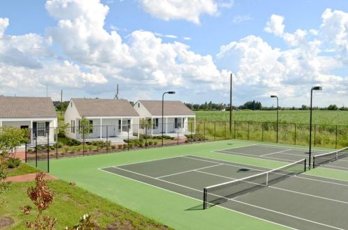 A view of tennis courts surrounded by green fields and modern white cottages under a partly cloudy sky.