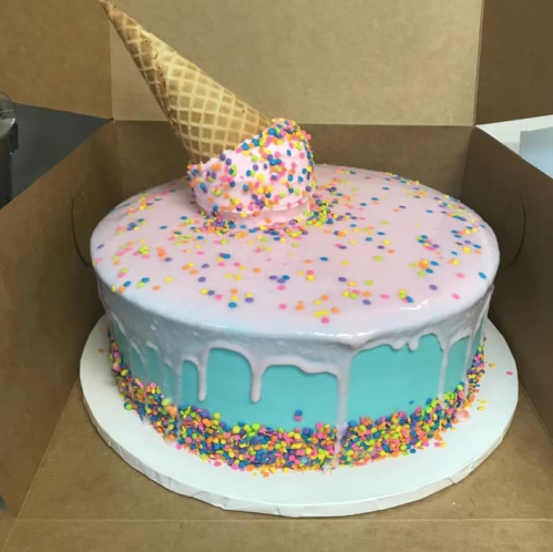 A colorful cake with pink and blue frosting, topped with a cone and sprinkles, in a brown box.