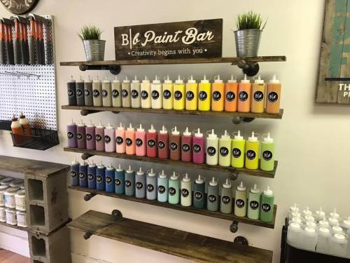 A wooden shelf displays colorful paint bottles at a paint bar, with a sign that reads "Creativity begins with you."