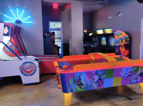 A colorful arcade with a neon light, featuring air hockey and various classic arcade games.
