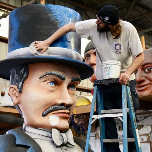 A person on a ladder paints a large, detailed statue of a man in a top hat, with other sculptures in the background.