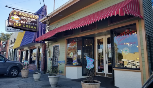 Colorful ice cream shop exterior with a striped awning and a neon sign, featuring vibrant window displays and potted plants.
