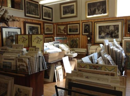 A cozy art gallery filled with framed paintings and stacks of prints displayed on tables and walls.