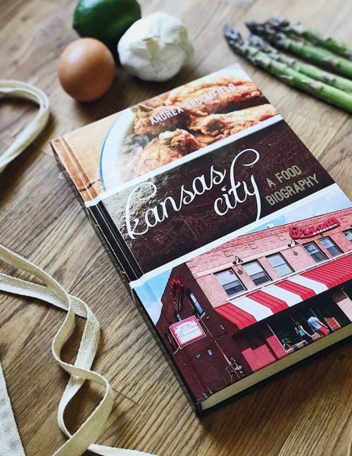 A food biography book titled "Kansas City" on a wooden surface, surrounded by fresh ingredients like asparagus, an egg, and garlic.
