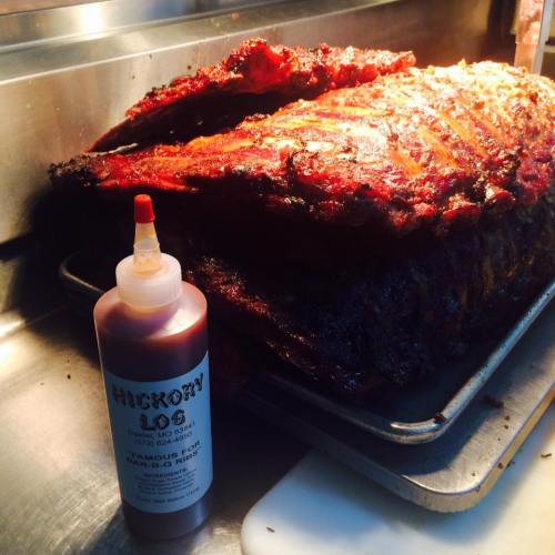 A tray of juicy, smoked ribs next to a bottle of Hickory Log barbecue sauce.