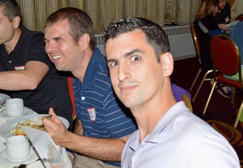Two men at a dining table, one smiling and eating, the other looking at the camera with a surprised expression.