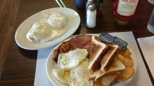 A plate of breakfast featuring eggs, ham, toast, chips, and a side of biscuits with gravy.