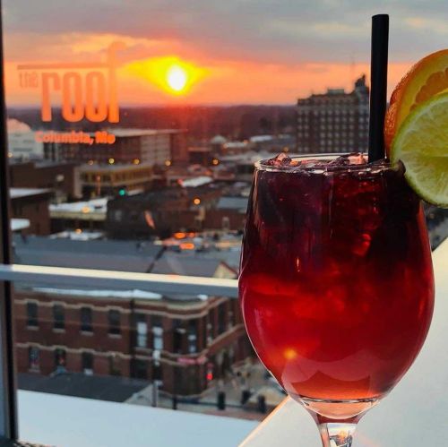 A vibrant cocktail with a slice of orange sits against a sunset view of Columbia, Missouri, from a rooftop bar.