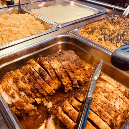 A buffet display featuring sliced meat, rice, and creamy dishes in metal trays. Tongs are placed beside the food.