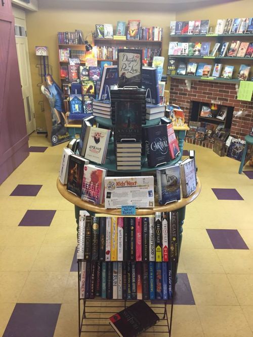 A colorful display of books on a cart in a cozy bookstore, surrounded by shelves filled with more books.