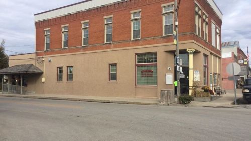 A two-story brick building with a beige facade, featuring large windows and a sign, located on a quiet street.