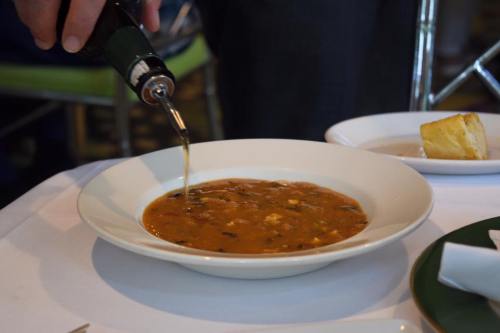 A person pours liquid into a bowl of soup on a white tablecloth, with a piece of bread on the side.