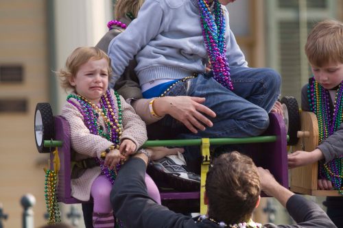 A child with a worried expression sits in a parade float, surrounded by others wearing colorful beads.