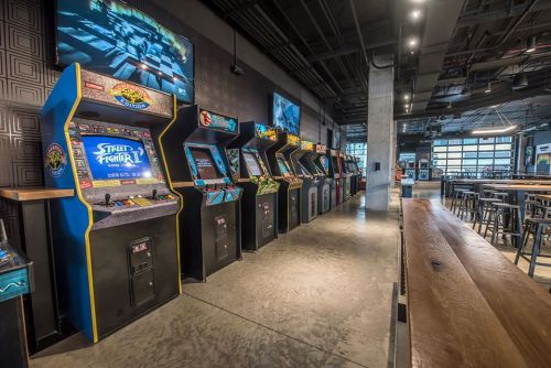 A row of vintage arcade machines in a modern gaming venue, with a bar and industrial decor in the background.