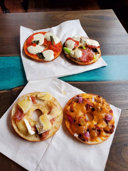 Four bagel halves topped with various ingredients: tomatoes, mozzarella, pepperoni, cheese, ham, and onions on a wooden table.