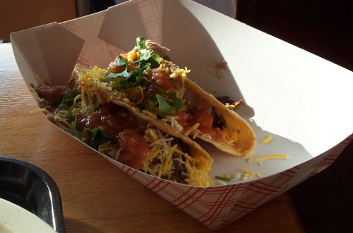 Three tacos in a paper tray, topped with cheese, lettuce, and salsa, sitting on a wooden table.