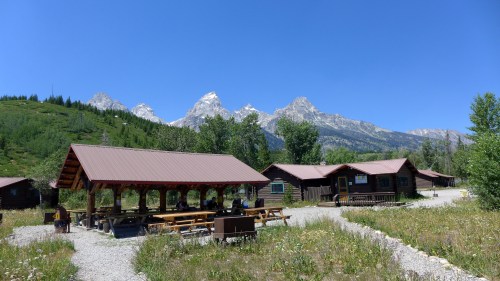 A rustic cabin area with picnic tables, surrounded by lush greenery and mountains under a clear blue sky.