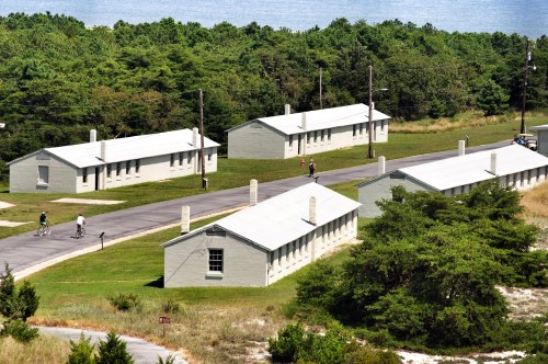 Aerial view of several light-colored buildings surrounded by greenery and a pathway, near a body of water.